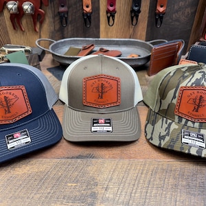 May include: Three trucker hats with a brown leather patch that says "Get Hooked" and a fish silhouette. The hats are in different colors and patterns: navy blue, green, and camouflage.