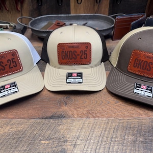 May include: Three trucker hats with a tan front panel and a mesh back. Each hat features a brown leather patch with the text "TENNESSEE GKOS-25 NASHVILLE". The hats have different colored mesh backs: white, black, and tan.