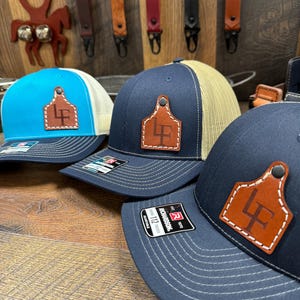May include: Three trucker hats with a brown leather patch featuring the initials "LF" on each. The hats are blue, turquoise, and beige. The hats are on a wooden surface.