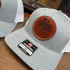 May include: Gray and white trucker hat with a brown leather patch featuring a tractor and the text "Matheny Farms". The hat has a Richardson 112 label.