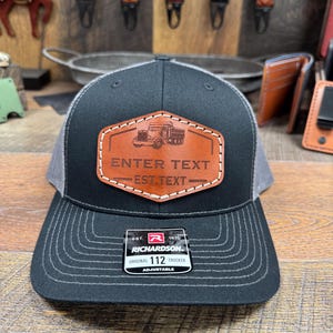 May include: Black and gray trucker hat with a leather patch featuring a truck graphic and the text "ENTER TEXT EST. TEXT". The hat is adjustable and has a Richardson 112 tag.