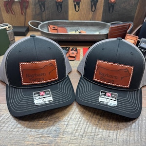 May include: Two black and grey trucker hats with brown leather patches that say "Rayborn Farms" and feature a silhouette of a cow. The hats are both Richardson brand.