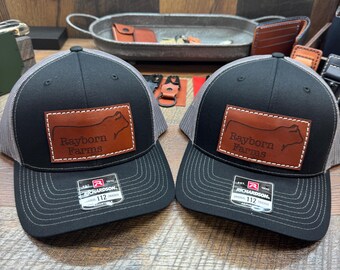 May include: Two black and grey trucker hats with brown leather patches that say "Rayborn Farms" and feature a silhouette of a cow. The hats are both Richardson brand.