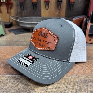 May include: Gray and white trucker hat with a brown leather patch featuring a tractor and the text "ENTER TEXT EST. TEXT".