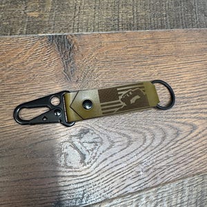 May include: A black leather keychain with a brown leather strap and a metal clasp. The strap has a design of an American flag with a fist in the center.