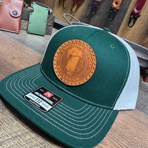 May include: A green and white trucker hat with a brown leather patch that says "Emotional Support Beverage". The patch features a beer mug with a foam head.