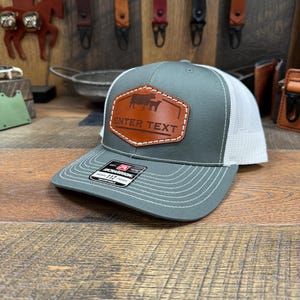 May include: A gray and white trucker hat with a brown leather patch featuring a cow and calf silhouette and the text "ENTER TEXT".