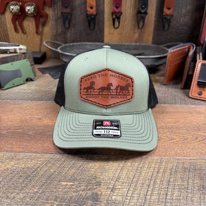 May include: A green and black trucker hat with a brown leather patch that says "Feed the Horses Shoot the Wolves". The patch features a silhouette of four horses running. The hat is adjustable and has a Richardson 112 label.
