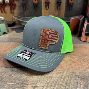 May include: A gray trucker hat with a neon green mesh back and brim. The hat features a leather patch with the word "POWERSTROKE" in a stylized design. The hat is adjustable and has green stitching.