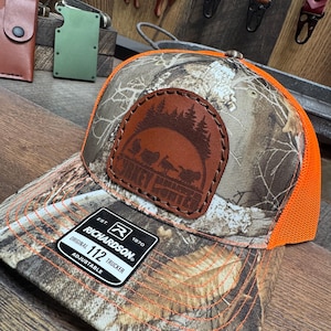 May include: A camouflage and orange trucker hat with a brown leather patch featuring a turkey and the text "Turkey Hunter". The hat is adjustable and has the Richardson brand logo and the text "Original 112 Trucker" on the side.