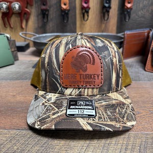 May include: A brown and tan camouflage trucker hat with a brown leather patch that says "Here Turkey Turkey Turkey". The hat is adjustable and has a Richardson 112 label.