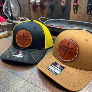 May include: Two baseball caps with a brown leather patch that says "REDEEMED" with a cross and a nail. One cap is black with a yellow mesh back and the other is brown with a brown mesh back.