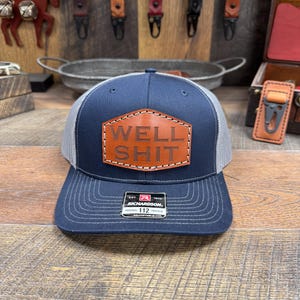May include: A navy blue and gray trucker hat with a brown leather patch that says "WELL SHIT" in white stitching. The hat is adjustable and has a Richardson 112 label.