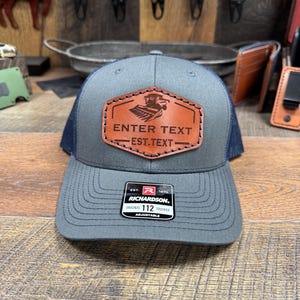 May include: A gray and blue trucker hat with a brown leather patch that says "ENTER TEXT - EST. TEXT". The patch features a silhouette of a combine harvester. The hat is adjustable and has a Richardson 112 label.