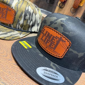 May include: Two snapback caps featuring camouflage patterns and leather patches. The front cap is black camouflage with a tan leather patch reading LINE LIFE IT'S A DIFFERENT BREED. The rear cap has a tan camouflage pattern and a similar patch.