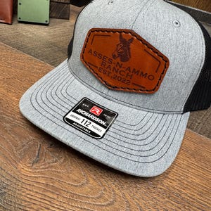 May include: Gray trucker hat with a black mesh back and a brown leather patch. The patch features a donkey graphic and the text "ASSES-N-AMMO RANCH EST.2022". The hat has a curved brim and an adjustable strap.