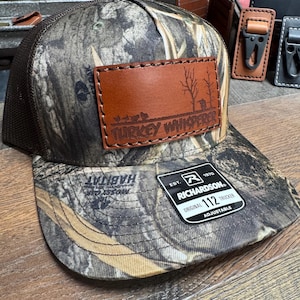 May include: A camouflage trucker hat with a brown leather patch that says "Turkey Whisperer". The hat is adjustable and has a black mesh back.