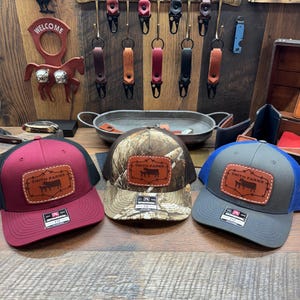 May include: Three trucker hats with a brown leather patch that says "Smith Farms" and a silhouette of a cow. The hats are red, camouflage, and gray.