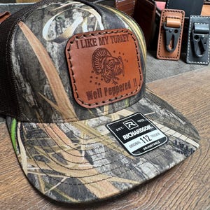 May include: A brown camouflage trucker hat with a brown leather patch that says "I Like My Turkey Well Peppered". The hat is adjustable and has a Richardson 112 logo.