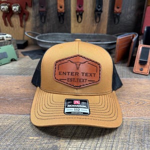 May include: A brown and black trucker hat with a brown leather patch that says "ENTER TEXT EST.TEXT" and a brown leather steer head design. The hat is adjustable and has a black mesh back.