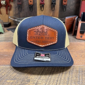 May include: A navy blue and tan trucker hat with a brown leather patch featuring a design of horses and the text "ENTER TEXT EST. TEXT". The hat has a Richardson 112 label.