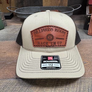 May include: A beige and black trucker hat with a brown leather patch that says "Billiards Room" with a pool cue and 8 ball graphic and "Rack 'Em Up" text. The hat has a Richardson logo and "Original 112 Trucker Adjustable" text.