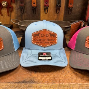 May include: Three trucker hats with a brown leather patch that says "Q-D Quarter Horse Est. 2019". The hats are blue, gray, and pink. The blue hat has a red and white Richardson label.