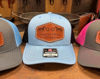 May include: Three trucker hats with a brown leather patch that says "Q-D Quarter Horse Est. 2019". The hats are blue, gray, and pink. The blue hat has a red and white Richardson label.