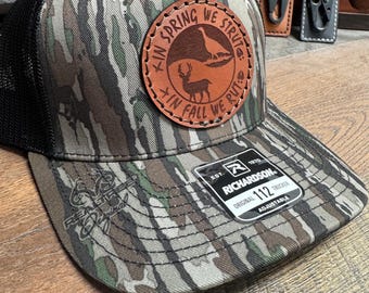 Puede incluir: Una gorra de camionero de camuflaje con un parche de cuero marrón que dice "Kin Spring We Strut: In Fall We Rut." La gorra es ajustable y tiene una parte trasera de malla negra.
