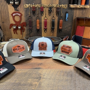 May include: Five different trucker hats with leather patches that say "PG and E" in a brown font. The hats are in different colors, including black, brown, green, blue, and gray.