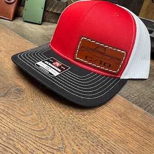 May include: A red and white trucker hat with a black brim. The hat features a brown leather patch with a truck design and the text "C/10". The hat is made by Richardson.