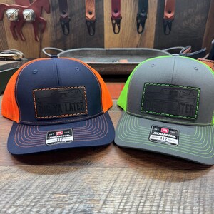 May include: Two trucker hats with a black patch that says "See Ya Later" in white lettering. The hat on the left is navy blue with an orange mesh back. The hat on the right is gray with a green mesh back.