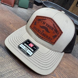 May include: Tan and brown trucker hat with a leather patch. The patch features the text "I'M YOUR HUCKLEBERRY" with a mustache and two crossed pistols. The hat is adjustable and has a Richardson 112 tag.
