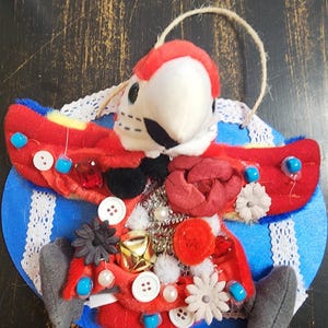 May include: A handmade parrot ornament with a red, white, and blue color scheme. The parrot has a plush white head and a red beak. The body is adorned with buttons, beads, and fabric flowers. The ornament is attached to a blue felt circle with white lace trim.