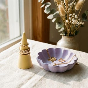 May include: A yellow cone-shaped ring holder displaying several gold-toned rings, positioned next to a lavender, scalloped-edge dish containing a gold-toned chain necklace and small stud earrings.