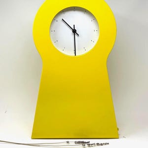 May include: A bright yellow keyhole-shaped clock with a white clock face. The clock has black hands and a minimalist design. The clock is a decorative item.