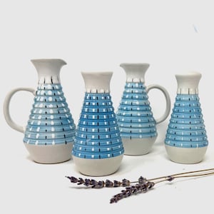 May include: Four ceramic vases with a blue and white dotted pattern. Two vases have handles. The vases have a light grey base and a white rim. A sprig of lavender is in the foreground.