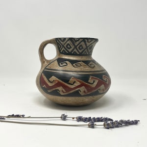 以下が含まれることがあります： 黒と赤の幾何学模様が施された、小さな茶色の陶器のピッチャー。ピッチャーは持ち手と丸いボディを持っています。