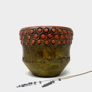 Peut inclure: Un pot en céramique à surface texturée. Le pot est principalement vert olive avec une bande de cercles orange près du sommet. Le bord est rouge. Le fond présente des lignes verticales. Quelques brins de lavande sont au premier plan.