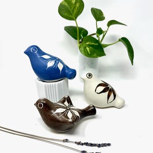 May include: Three ceramic bird figurines in blue, white, and brown, each with a white floral design. The birds are arranged on a white surface with a small potted plant and lavender sprigs.