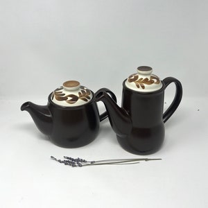 May include: Two brown ceramic teapots with white lids and brown floral designs. The teapots have a brown glaze and are sitting on a white surface.