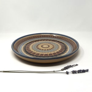 May include: A large, round ceramic serving platter with a brown, blue, and white geometric design. The design features concentric circles and a sunburst pattern.
