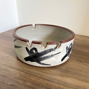 May include: Ceramic ashtray with a cream-colored interior and exterior. The rim is brown, and the exterior features black abstract brushstrokes. The ashtray has four cigarette rests.