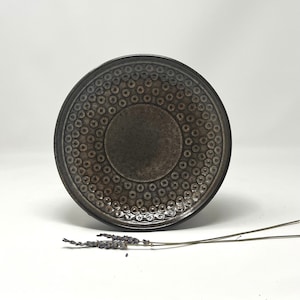 May include: A dark brown ceramic plate with a raised circular pattern. The plate has a slightly textured surface.