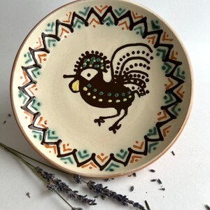 May include: A ceramic plate with a brown rooster design. The rooster has a yellow beak, green and yellow accents on its head, and a brown tail with white dots. The plate has a brown, orange, and green geometric border.