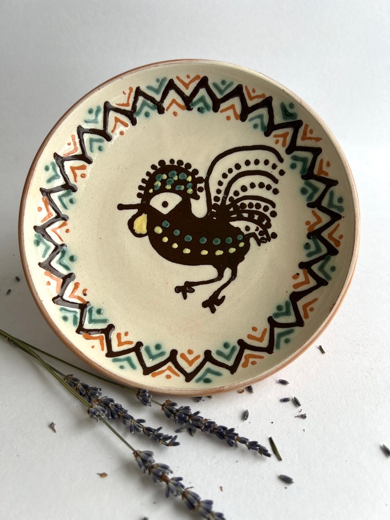 May include: A ceramic plate with a brown rooster design. The rooster has a yellow beak, green and yellow accents on its head, and a brown tail with white dots. The plate has a brown, orange, and green geometric border.