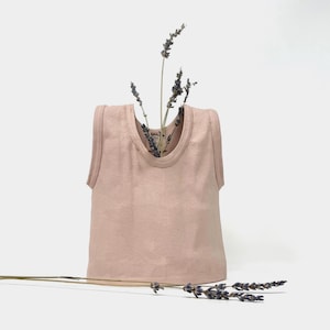 May include: A light pink tank top with a round neckline. The tank top is displayed with sprigs of lavender. The fabric has a ribbed texture. The overall aesthetic is minimalist and natural.