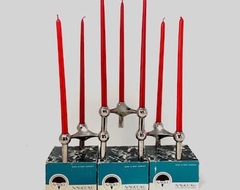 Puede incluir: Un sistema de candelabros cromados modulares compuesto por tres unidades metálicas interconectadas, cada una sobre una caja azul y negra con la etiqueta Nagel KG Wesseling-Köln. Seis velas cónicas rojas están insertadas en los soportes.