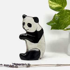 May include: A decorative panda figurine with black and white coloring. The panda is in a sitting position with its arms raised. The panda's body has a textured pattern. The figurine is on a white surface.