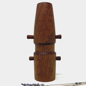 以下が含まれることがあります： ユニークなレイヤードデザインの手作りの木製花瓶。花瓶は、豊かで濃い色の木材で作られており、円筒形のベースとフレアトップが特徴です。小さな水平木製ハンドルが側面に付いています。乾燥したラベンダーの小枝が前景にあります。